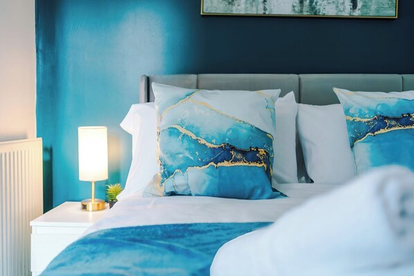 A bed with a white headboard, two pillows with blue and gold patterns, a white bedside table with a lamp, and a blue wall.