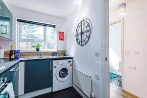 A compact kitchen with a built-in washing machine, a window with blinds, and a wall-mounted clock.