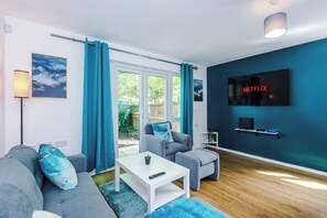 A modern living room with a grey sofa, a white coffee table, and a grey armchair. A TV mounted on the wall displays the Netflix logo.