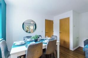 Un coin repas avec une table blanche dressée pour quatre personnes, entourée de chaises, un miroir rond accroché au mur et des portes en bois.