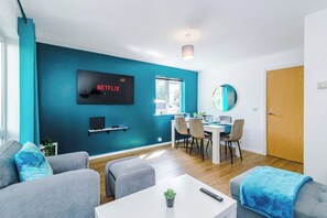 Un salon moderne avec un mur d’accent de couleur turquoise, un téléviseur à écran plat, un coin repas avec une table et des chaises, et une porte en bois.