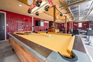A pool table with billiard balls and cues, a red wall with a wooden panel, and a ceiling with exposed beams and lighting fixtures.