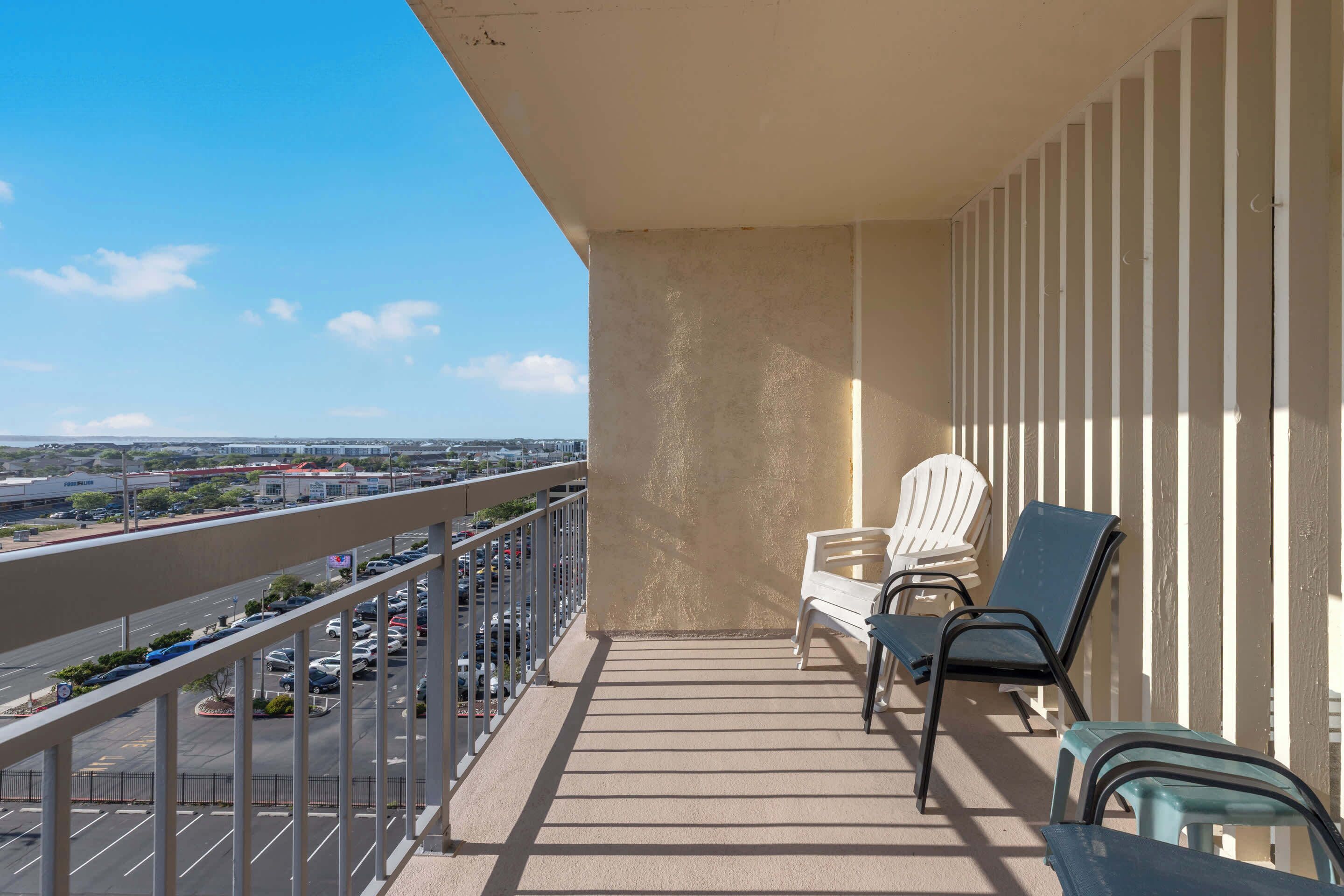 Un balcon donnant sur un parking et sur des bâtiments éloignés, une chaise blanche, une chaise bleue et une petite table.