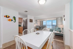 Eine moderne Küche mit weißen Schränken, ein Essbereich mit einem weißen Tisch und Stühlen sowie ein Wohnzimmer mit einer Couch und Meerblick.