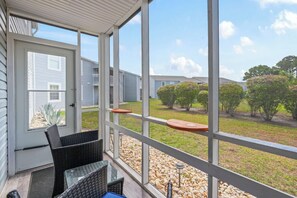 Eine überdachte Veranda mit Korbmöbeln, einer Tür zum Garten und Blick auf Nachbarhäuser und Grünflächen.