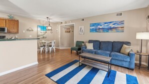 Ein modernes Wohnzimmer mit einem blauen Sofa, einem Couchtisch, einem Essbereich mit weißem Tisch und Stühlen sowie einer Küchenzeile mit Holzschränken und einer Mikrowelle.