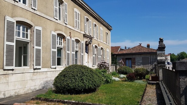 Ein historisches Gebäude mit Rundbogenfenstern und Fensterläden, ein gepflegter Garten und ein traditionelles Ziegeldach.