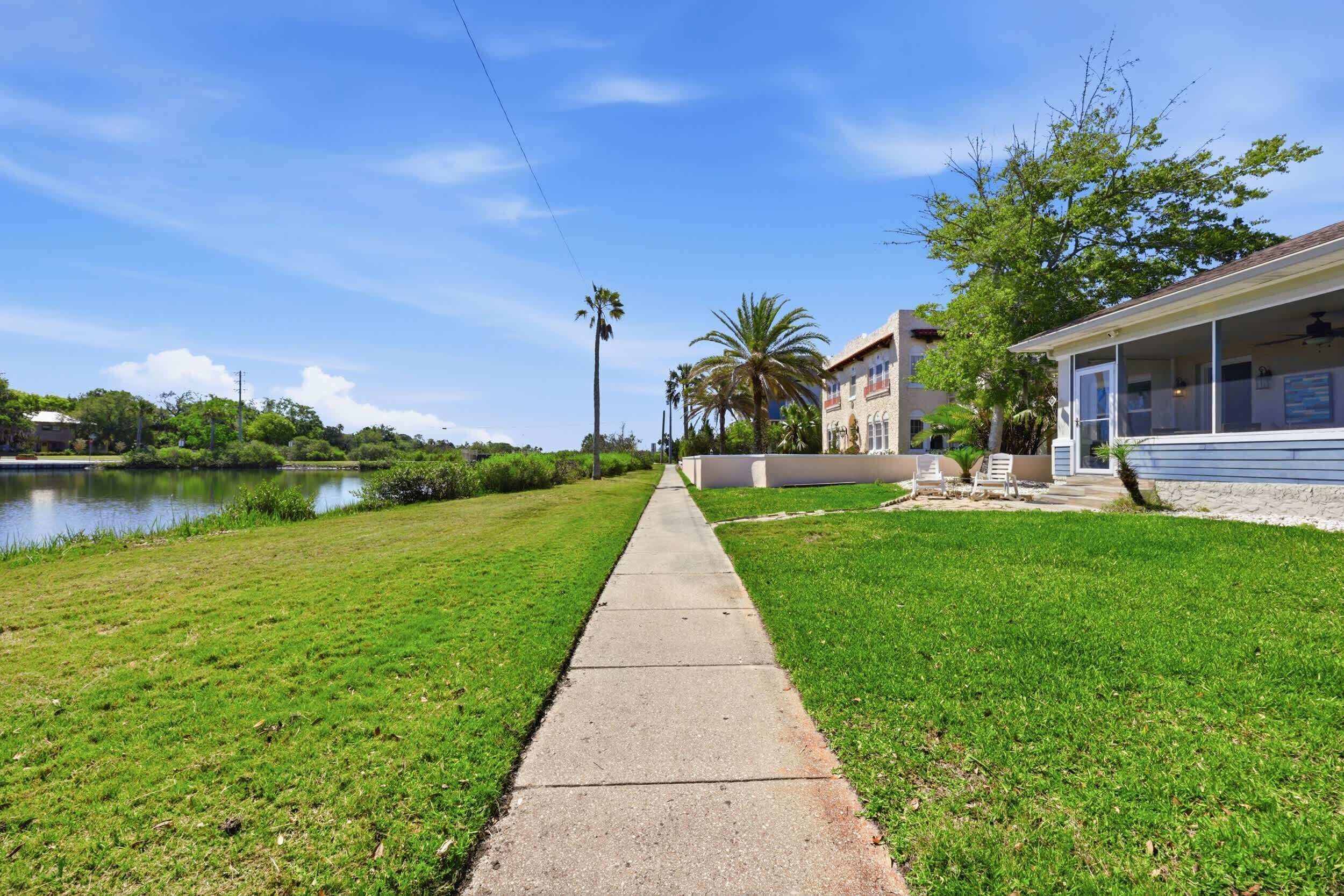 A pathway leading to a building with a pool, surrounded by greenery and a clear sky.
