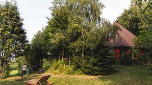 A wooden deck chair in a grassy area with trees and a red-roofed building in the background.