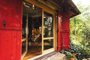 A red wooden cabin with a glass door leading to an interior with a hammock and a chair. - Bukowica Miła - Nature Escape with eco sauna (Wola Piotrowa)