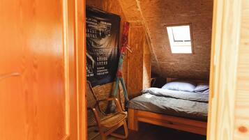 Een kleine, rustieke kamer met een houten plafond, een enkel bed met een beddegoed met dessin, een rieten stoel en een schoolbord met tekst erop.