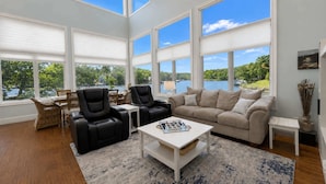 A spacious living room with large windows, a beige sofa, black chairs, a white coffee table, and a view of a lake. - Lake Paradise Lakefront Stay (Eldon)