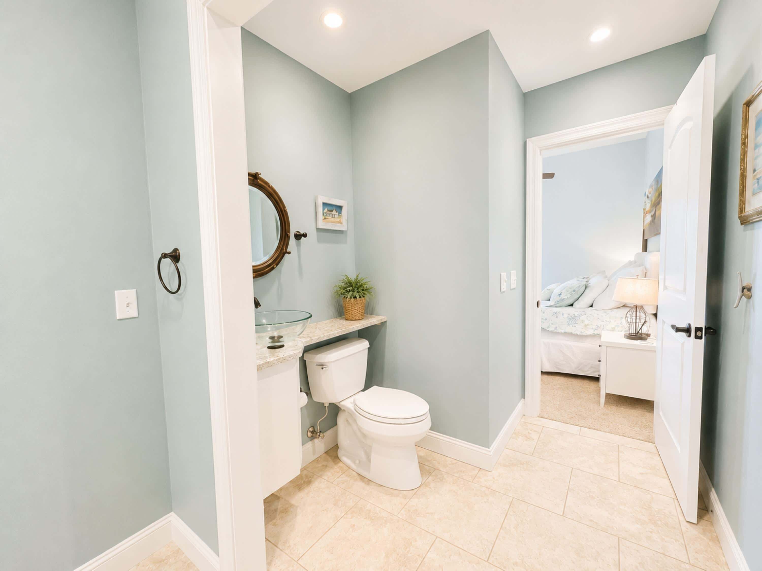 A bathroom with a toilet, sink, and mirror, leading to a bedroom with a bed and nightstand.