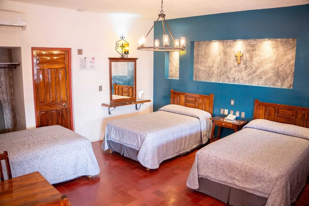 Habitación de hotel con tres camas, una puerta de madera, un espejo, una araña y un aplique de pared.