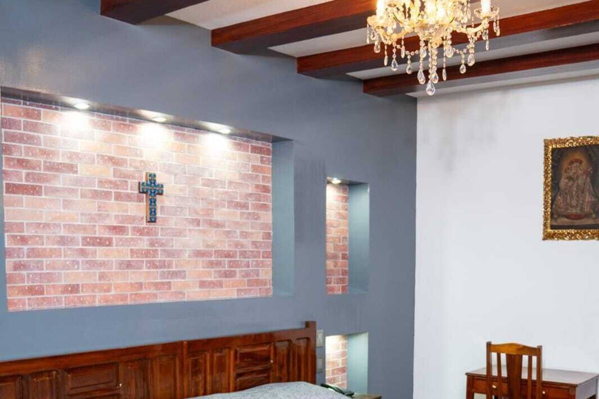 Un dormitorio con una cama grande, un cabecero de madera, una pared de ladrillo con una cruz, una araña y una pequeña mesa con una silla.
