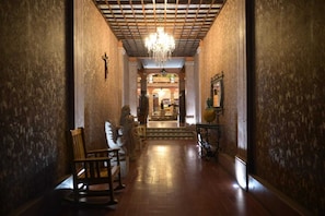 A hallway with a chandelier, wooden chairs, and a cross on the wall.