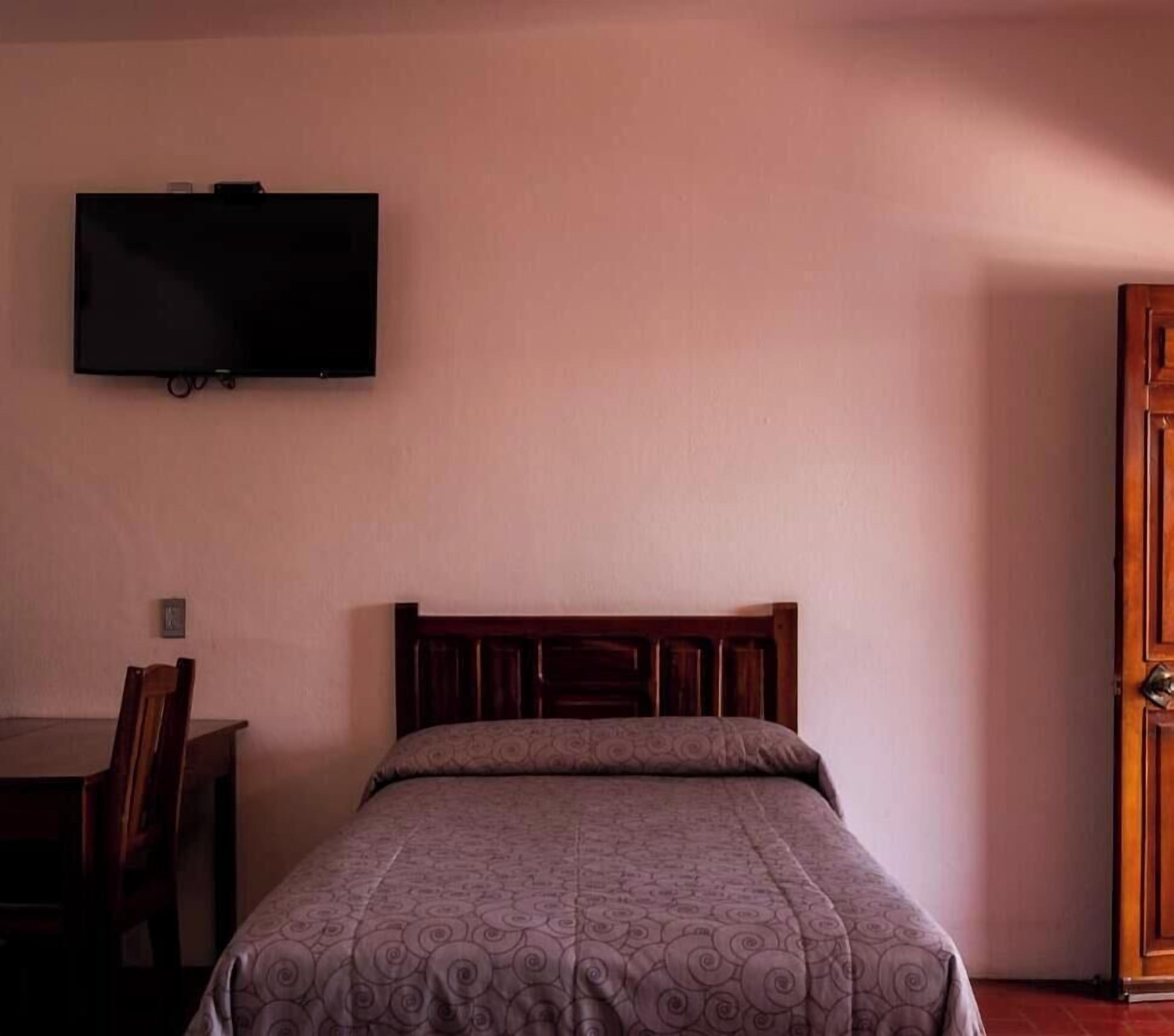 A single bed with a wooden headboard, a television mounted on the wall, a wooden door, and a desk with a chair.