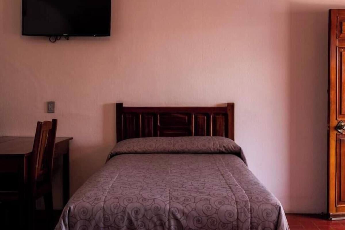 Una cama individual con cabecera de madera, un televisor montado en la pared, una puerta de madera y un escritorio con una silla.