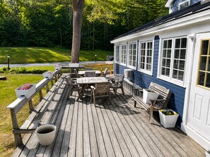 Eine Holzterrasse mit Gartenmöbeln, einer weißen Mülltonne und einem Gebäude mit großen Fenstern.