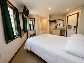 Une chambre à coucher avec un lit, un téléviseur fixé au mur, une salle de bain avec douche, et une porte donnant sur une autre pièce.