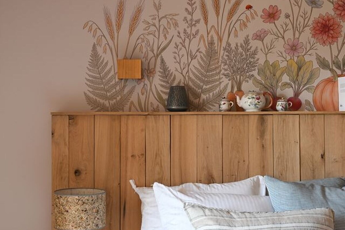 Cabeceira de madeira com papel de parede estampado com flores, uma luminária e uma cama com almofadas.
