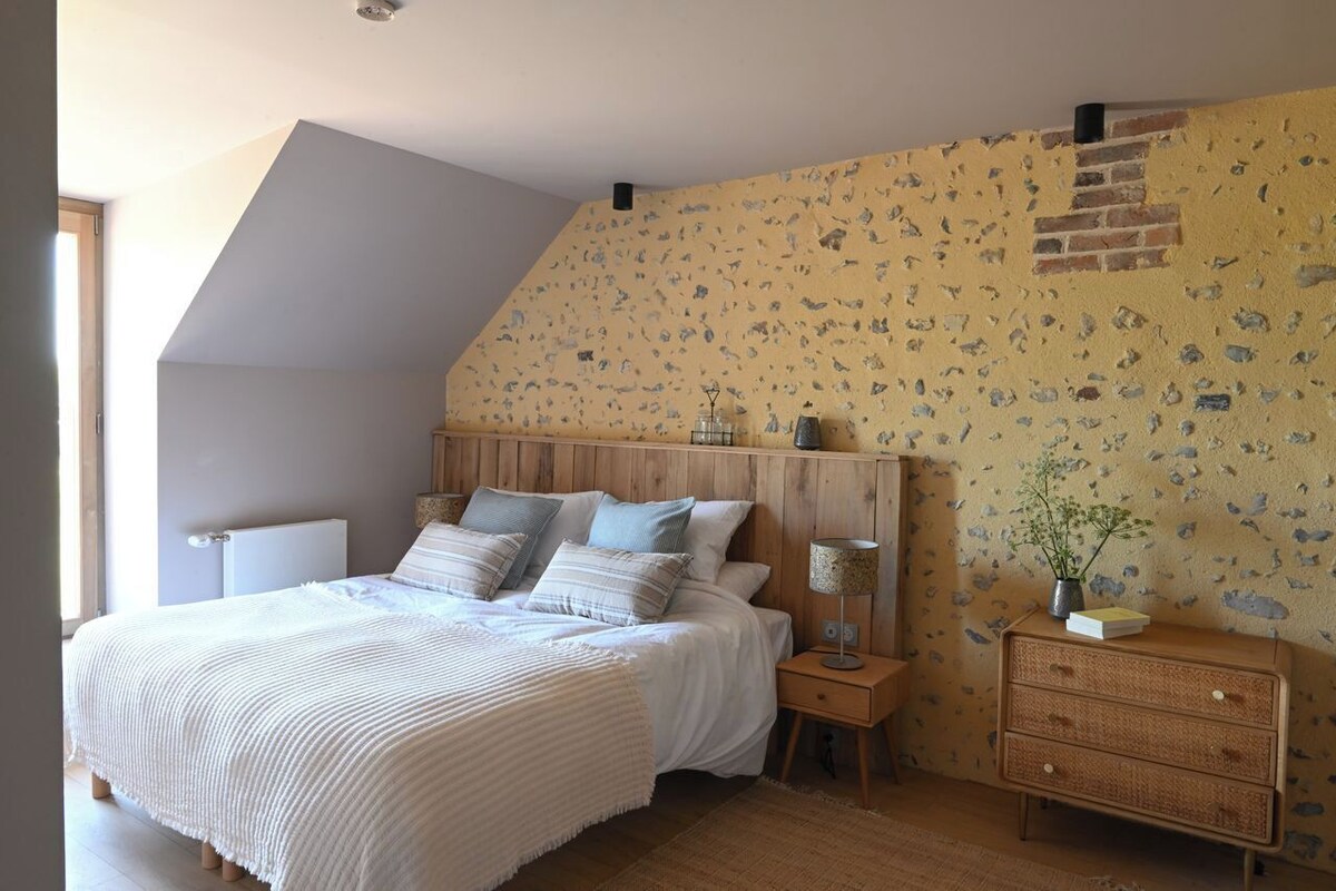 Quarto com teto inclinado, cama com várias almofadas, mesa de cabeceira com abajur e cômoda.