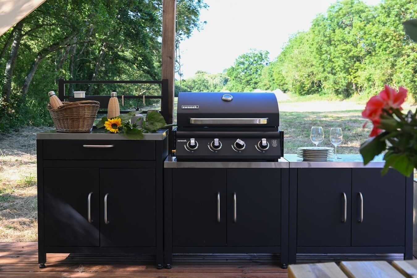A black outdoor grill with four control knobs, a basket, a vase with a sunflower, and a table with plates and glasses.