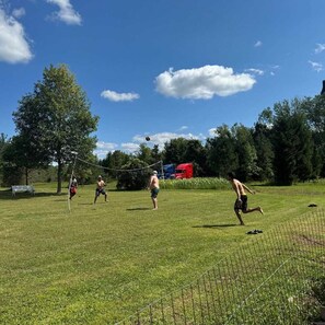 Un champ d’herbe avec un filet de volley-ball, des arbres et un camion rouge en arrière-plan.