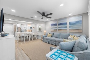 A modern living room with a grey sofa, a dining area with white chairs, and a kitchen in the background. - Sugar Beach F-28 ~ Gulf-side luxury townhome! (Navarre)