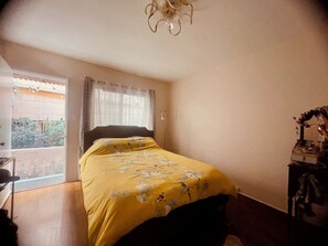 A bedroom with a bed, a window with curtains, a door, and a small table with items on it. - Spacious, Coastal 1BR Condo - Walk to Beach, Dining & Shopping in DT Long Beach! (Long Beach)