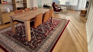 Ein modernes Wohnzimmer mit Esstisch aus Holz, bequemen Sitzgelegenheiten, einem Bücherregal und einem großen Fenster mit transparenten Vorhängen.