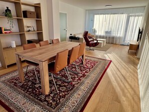 Ein modernes Wohnzimmer mit Esstisch aus Holz, bequemen Sitzgelegenheiten, einem Bücherregal und einem großen Fenster mit transparenten Vorhängen.