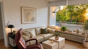 Ein modernes Wohnzimmer mit Ledersessel, Sofa, Couchtisch, Beistelltisch mit Lampe, gerahmtem Bild, Fenster mit Blick auf Bäume und Heizkörper.