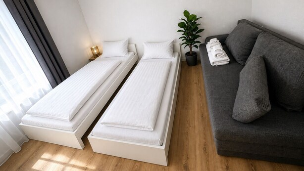 Ein Zimmer mit zwei Einzelbetten, einem grauen Sofa, einer Topfpflanze und einem Fenster mit Vorhängen.