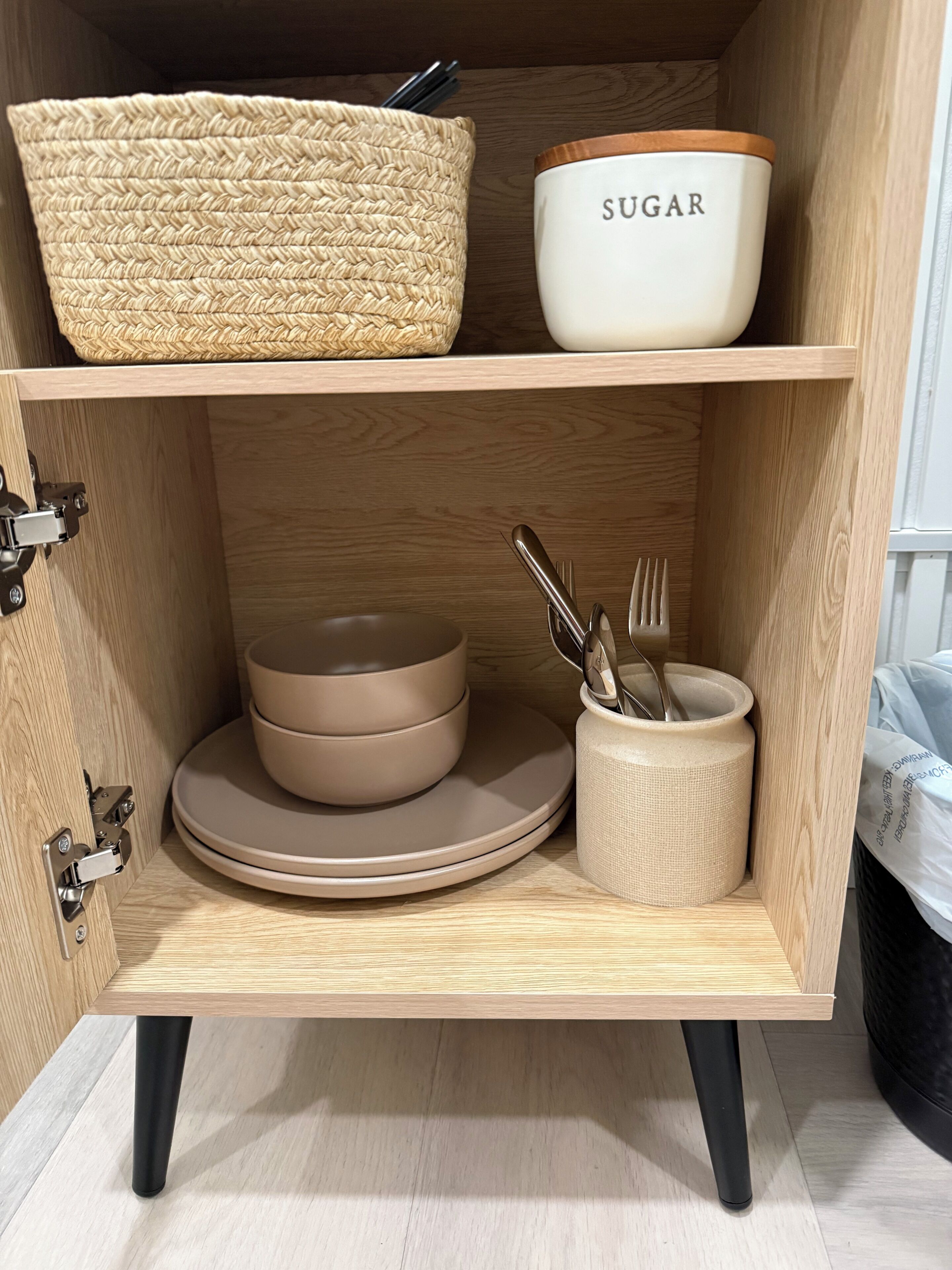 Un meuble en bois doté d’une porte à charnières, contenant un panier tressé, un récipient à sucre, des assiettes empilées et un porte-ustensiles.