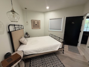 A bedroom with a bed, a bedside table, a lamp, a framed picture, a window with blinds, and a door.
