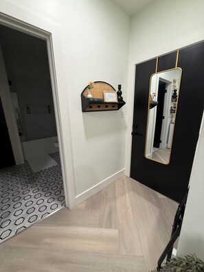 Un pasillo con suelo a cuadrícula blanco y negro, una pared negra con un espejo de marco dorado y un estante de madera con objetos decorativos.