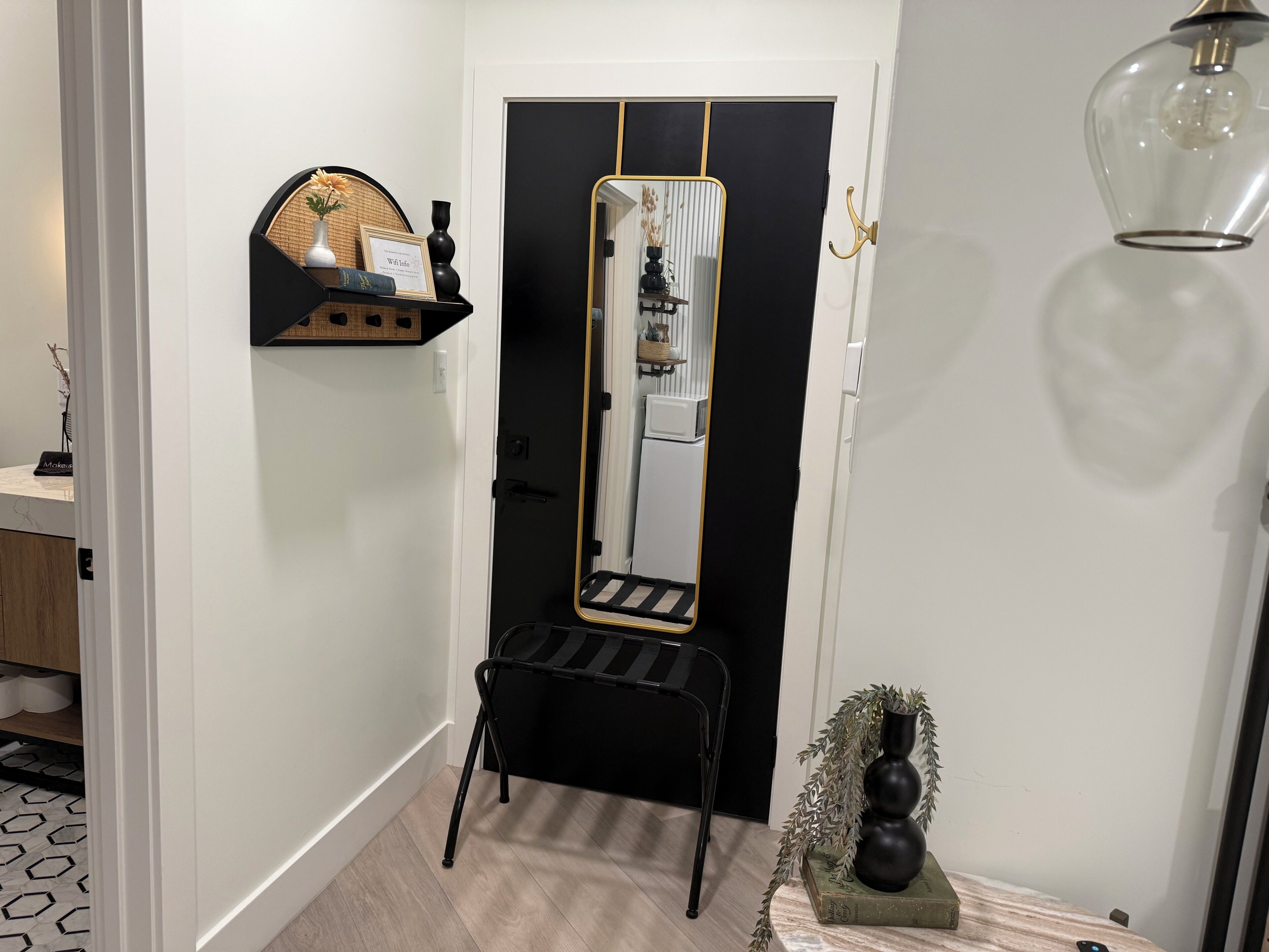 Un couloir avec un miroir à cadre noir, une chaise noire et une petite table sur laquelle se trouve un vase.