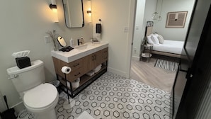 Ein modernes Badezimmer mit Toilette, Waschbecken und Spiegel. Ein Bett ist durch eine offene Tür im angrenzenden Zimmer sichtbar.