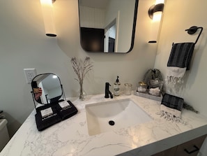 A bathroom with a white marble sink, a mirror, a black towel rack, a vase with dried plants, and a bottle of hand soap.