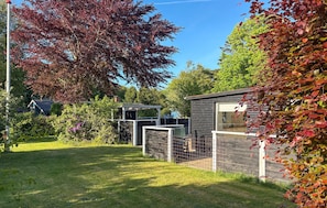 Ein von einer Hecke umgebener Gartenbereich mit einer kleinen Holzkonstruktion, einem Metalltor und einem Baum mit roten Blättern.