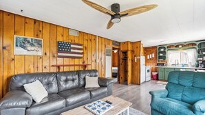 A cabin interior with a leather sofa, a blue armchair, a wooden coffee table, a ceiling fan, and a kitchen area with a refrigerator and microwave. - #2 | Cozy & Easy - Steps to the Sand (Oscoda)
