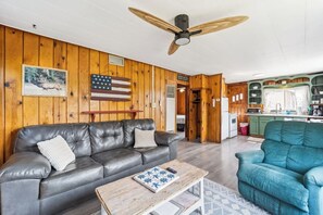 A cabin interior with a leather sofa, a blue armchair, a wooden coffee table, a ceiling fan, and a kitchen area with a refrigerator and microwave.
