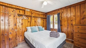 A wooden-paneled bedroom with a bed, a dresser, a ceiling fan, and a window with curtains. - #2 | Cozy & Easy - Steps to the Sand (Oscoda)
