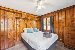 A wooden-paneled bedroom with a bed, a dresser, a ceiling fan, and a window with curtains.