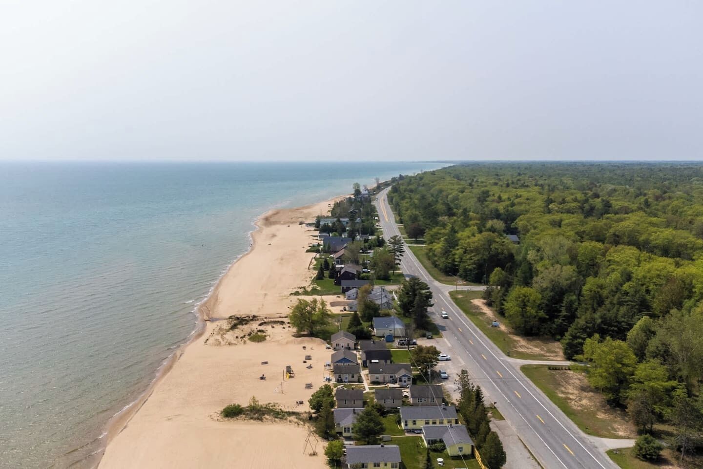 An aerial view of a coastal residential area with houses along the beach, a road, and dense forest.