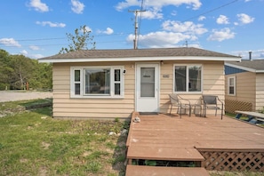 A single-story mobile home with a front door, two windows, a small deck with a table and chairs, and a visible utility pole.