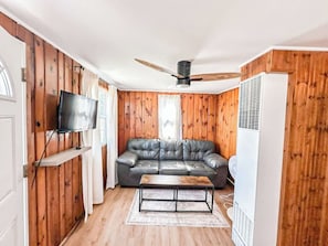 Una habitación con paredes de madera, un sofá de cuero, un televisor montado en la pared, un ventilador de techo y un refrigerador blanco.