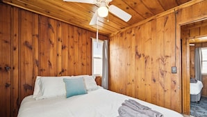 Habitación con paneles de madera, una cama, un ventilador de techo y vista a otra habitación.