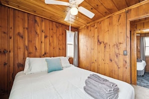 A wooden-paneled room with a bed, a ceiling fan, and a view of another room.
